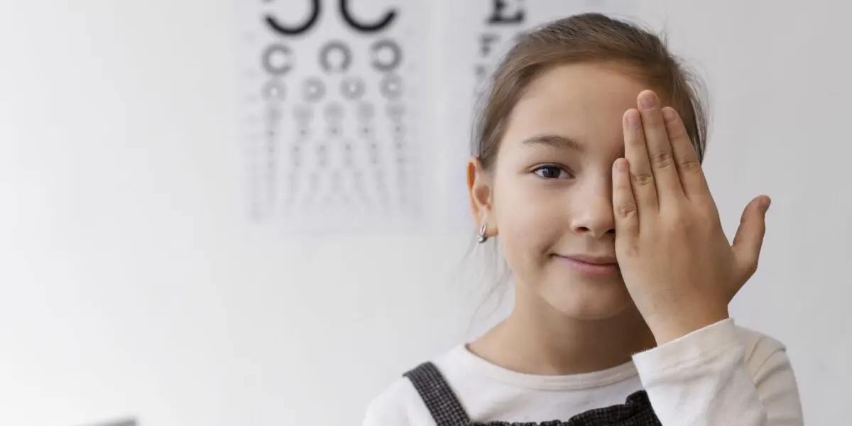 Smiling young girl covering one eye during a vision test at an eye clinic, with eye charts visible in the background