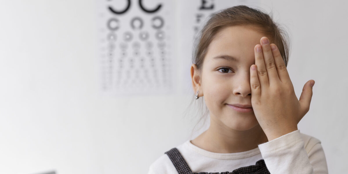 Smiling young girl covering one eye during a vision test at an eye clinic, with eye charts visible in the background